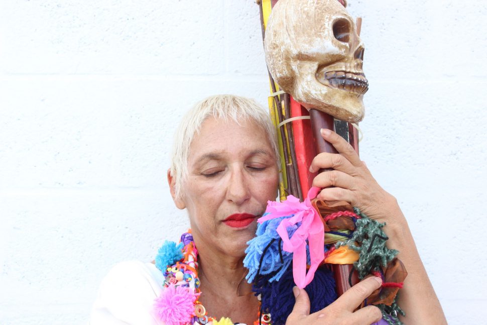 Close-up of the lead artist who wears bright red lipstick and a vibrant necklace. They also hold a sculptural object with colored streamers and a skull at the top.