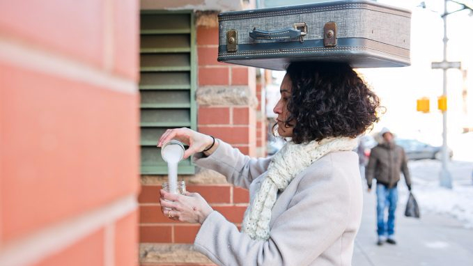 Lead project artist wears a scarf and coat while balancing a suitcase on their head. They are pouring a white, salt-like substance between two mason jars.