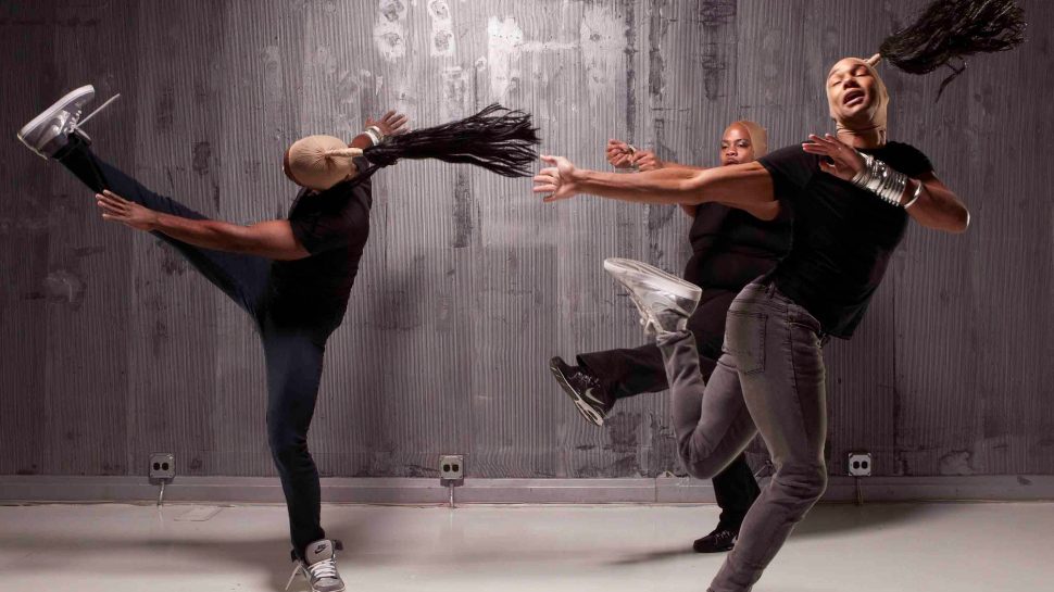 Three dancers kick, run, and spiral in jeans, black t-shirts, and high top sneakers. Two have special hairpieces that create long, flying ponytails.