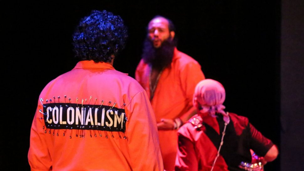 Performer in the foreground wears an orange jumpsuit with a large black patch attached with many safety pins. 